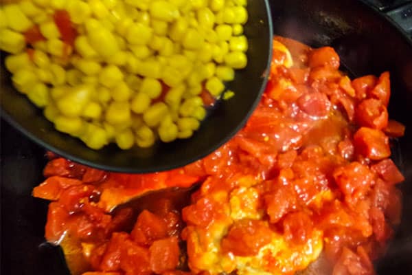 corn and peppers being added into tomatoes and chicken in a skillet