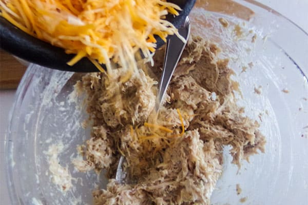 cheese being added to shredded chicken and cream cheese in a glass bowl