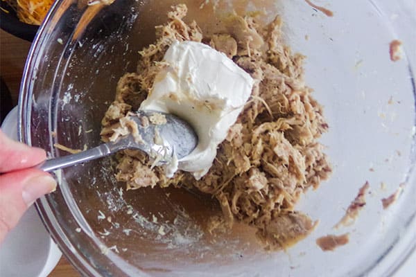 using a spoon to mix cream cheese and shredded chicken in a glass bowl
