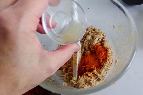 a hand pouring lime juice into a glass bowl of shredded chicken and seasonings