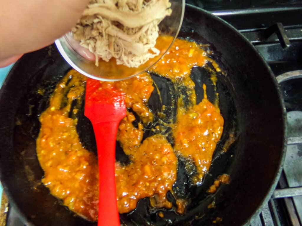 shredded chicken being added to tomato sauce and seasoning in a skillet