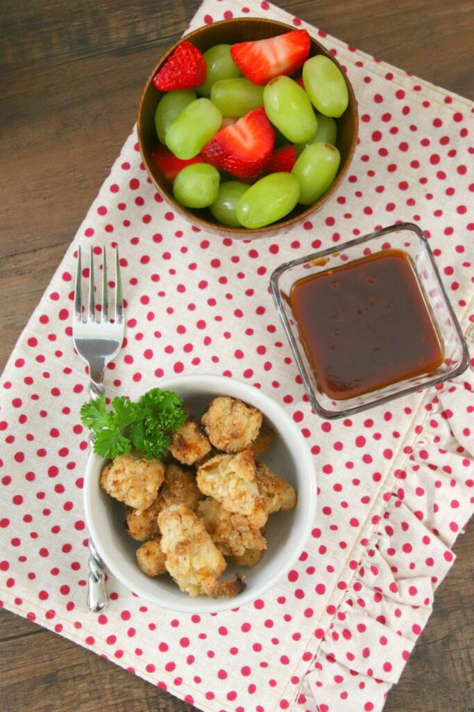 Air Fryer Chicken Bites in a bowl next to  sauce in a square bowl and fruits in another bowl, all on a white cloth with red polka dots. 