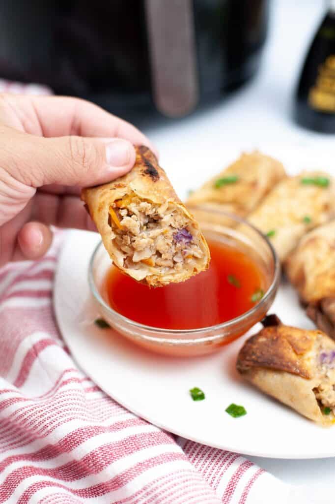 a hand holding half of an egg roll above more Air Fryer Spring Rolls with dipping sauce on a plate and garnished with green onion 