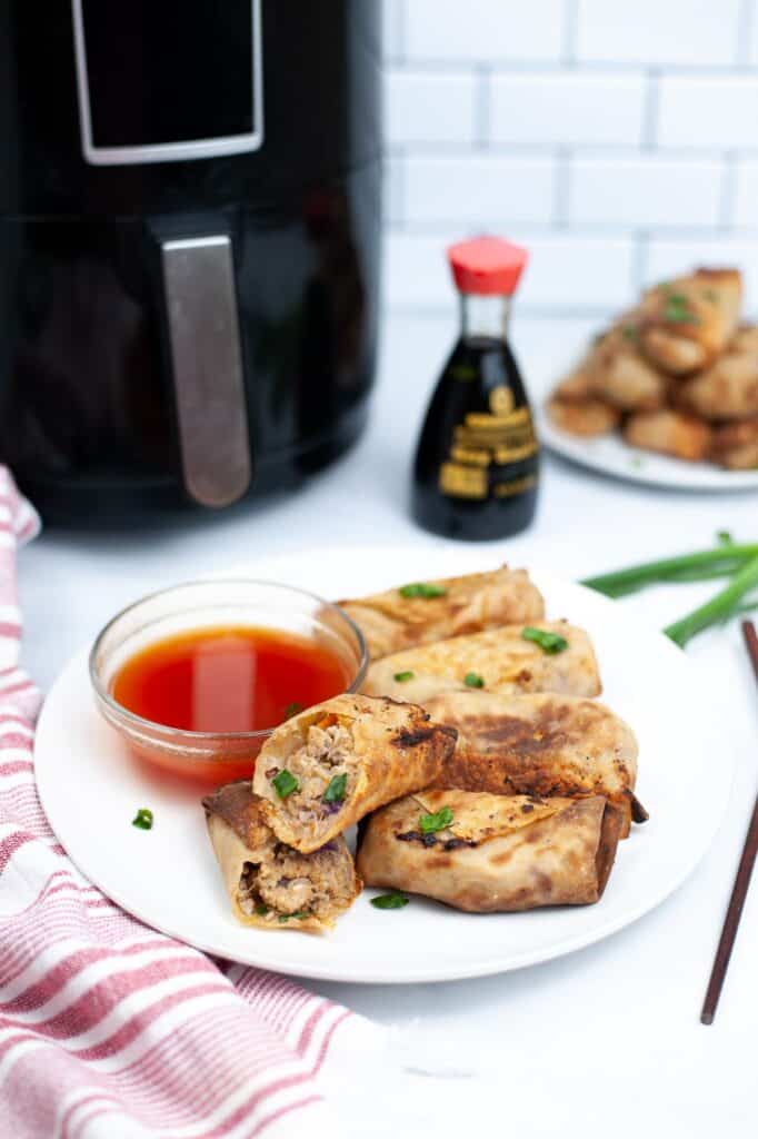 Air Fryer Spring Rolls with dipping sauce on a plate and garnished with green onion with an air fryer, soy sauce, and more spring rolls on a plate in the background