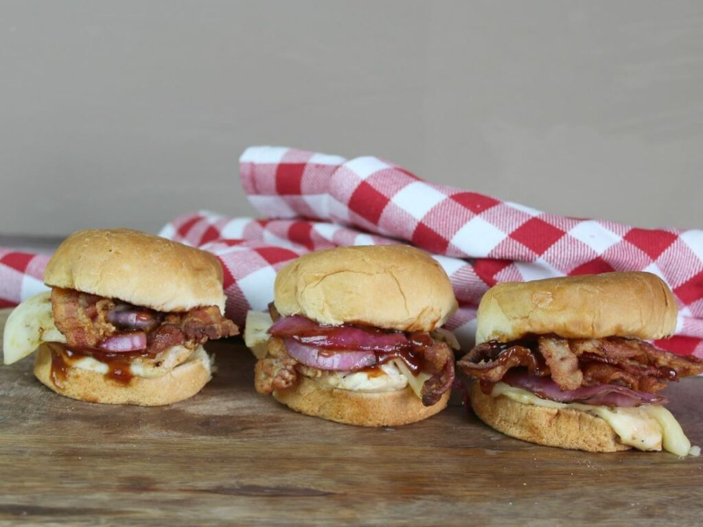 three barbecue chicken sliders on a wood table with a red and white checkered cloth behind them.