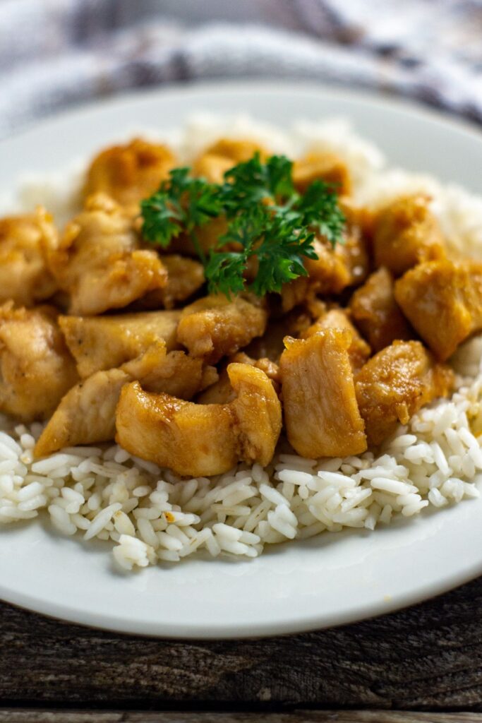 a closeup view of Honey Garlic Chicken on top of rice on a white plate next to a brown and white striped cloth
