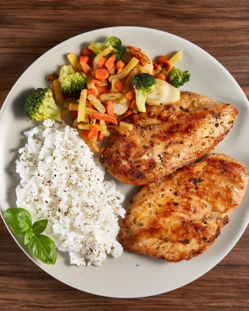 2 chicken breasts next to vegetables and rice on a white plate.