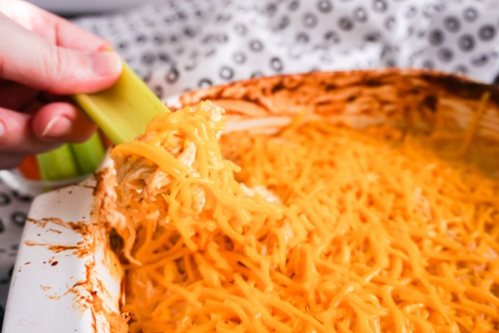 Hand holding a celery stick with buffalo chicken dip on it above dip in a dish.